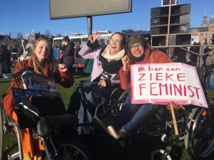 Foto van de Women's March. Naomi Doevendans, Annika Mell en Tamar Doorduin zitten met z'n drieen naast elkaar in hun rolstoelen, hun ogen dichtknijpend tegen de zon. Annika maakt een peace-teken, Tamar en Naomi heffen hun vuist. Tamar houdt een protestbord vast met "Ik ben een zieke feminist" erop, in grote roze letters.