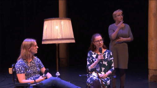 Tamar (links) en Eline (rechts) zitten op een podium, Tamar in haar rolstoel met de beensteunen omhoog, Eline op een stoel, haar stok tegen haar schoot aan geleund. Rechts achter Eline staat de gebarentolk. Tamar heeft schouderlang middenblond haar en draagt een bril, een donkerblauw bloemetjesshirt en een spijkerbroek. Eline heeft lang, half opgestoken kastanjebruin haar, een bruine bril en draagt een jurk met een grafisch patroon in wit en donkerblauw.