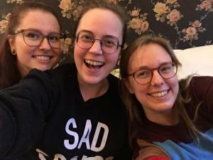 Selfie met van links naar rechts Eline, Annika en Tamar voor een vintage rozenbehangetje (donkerblauwe achtergrond met lichtroze rozen). We zien er zo uit: drie vrouwen met alle drie een bril op. Eline heeft half opgestoken kastanjebruin haar, Annika heeft donkerblond haar in een staart en een trui met "SAD" erop en Tamar heeft middenblond los haar.
