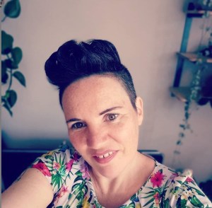 Foto van Xandra Koster. Xandra heeft een witte huidskleur en kort donkerbruin haar, opgeschoren aan de zijkanten en met een grote kuif bovenop. Ze glimlacht stoer maar ook enigszins bescheiden in de camera. Ze draagt een bloemetjesshirt in heldere kleuren. Op de achtergrand zijn kamerplanten te zien.