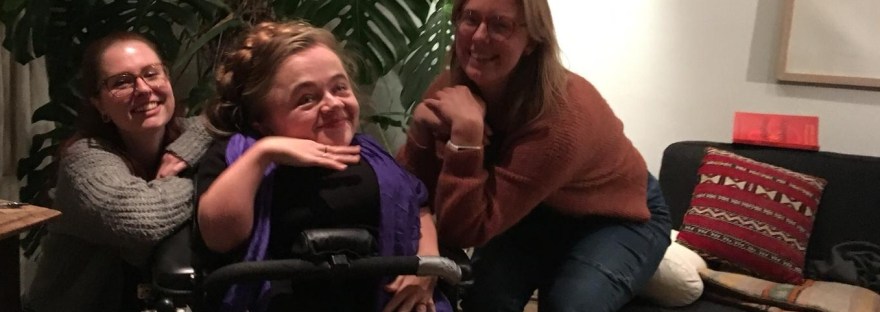 Eline, Mira en Tamar voor een grote kamerplant. Eline en Tamar leunen op de armleuningen van Mira's rolstoel en Mira houdt een hand onder haar kin.