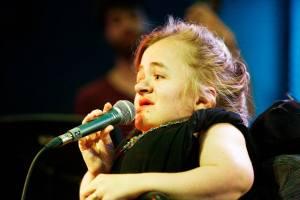 Foto van Mira tijdens een optreden. Ze draagt iets zwarts zonder mouwen, heeft rode lippenstift op en houdt een microfoon vast.