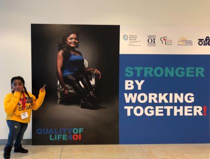 Chedda op een congres van Stichting Brittle Bones, onder een levensgrote campagneposter met haar erop. Chedda heeft lang, zwart haar, een bruine huidskleur, en ze is klein. Op de poster zien we Chedda in een zwarte leren broek en een glimmende, koningsblauwe top, zittend op een zilveren stoel. De Chedda die naast haar poster staat heeft haar hand voor de mond geslagen en wijst naar de poster, ze heeft haar haar in twee vlechten en draagt een gele Dipsaus trui. Naast de poster staat in grote letters "Stronger by working together".