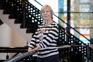 Foto van Margriet, een witte vrouw van middelbare leeftijd met kort lichtblond/grijs haar, die met haar niet-normatief gevormde handen een trapleuning vasthoudt